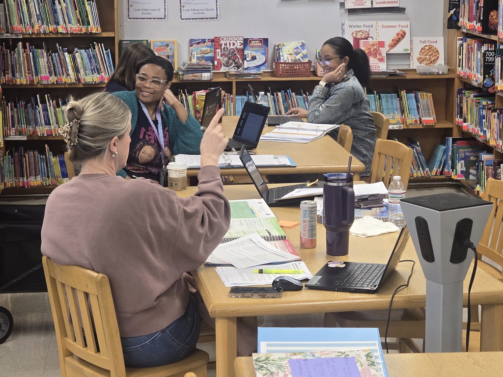 Four adults in a library, seated around a table, engaged in a discussion. They have laptops and tablets in front of them.