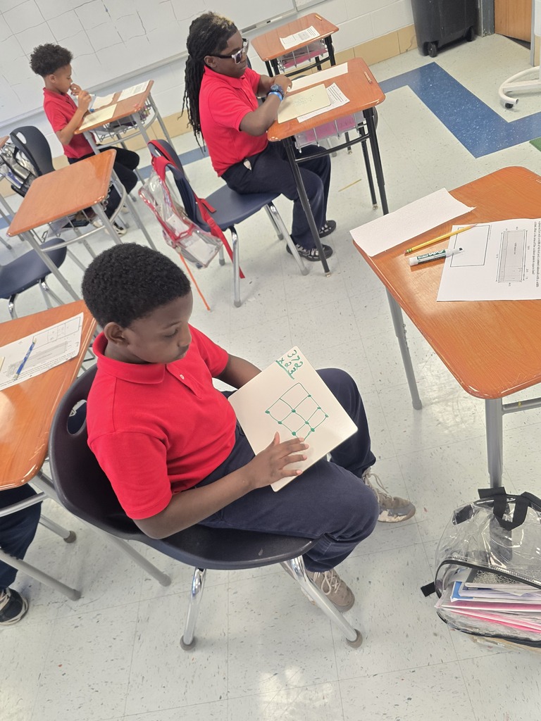 Students in red shirts sit at desks, focused on worksheets, with a blue carpeted floor.