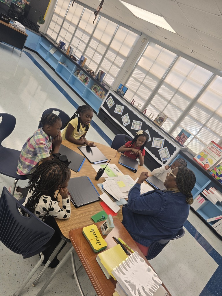 Four children and a teacher sit around a table in a classroom, working on papers and books.