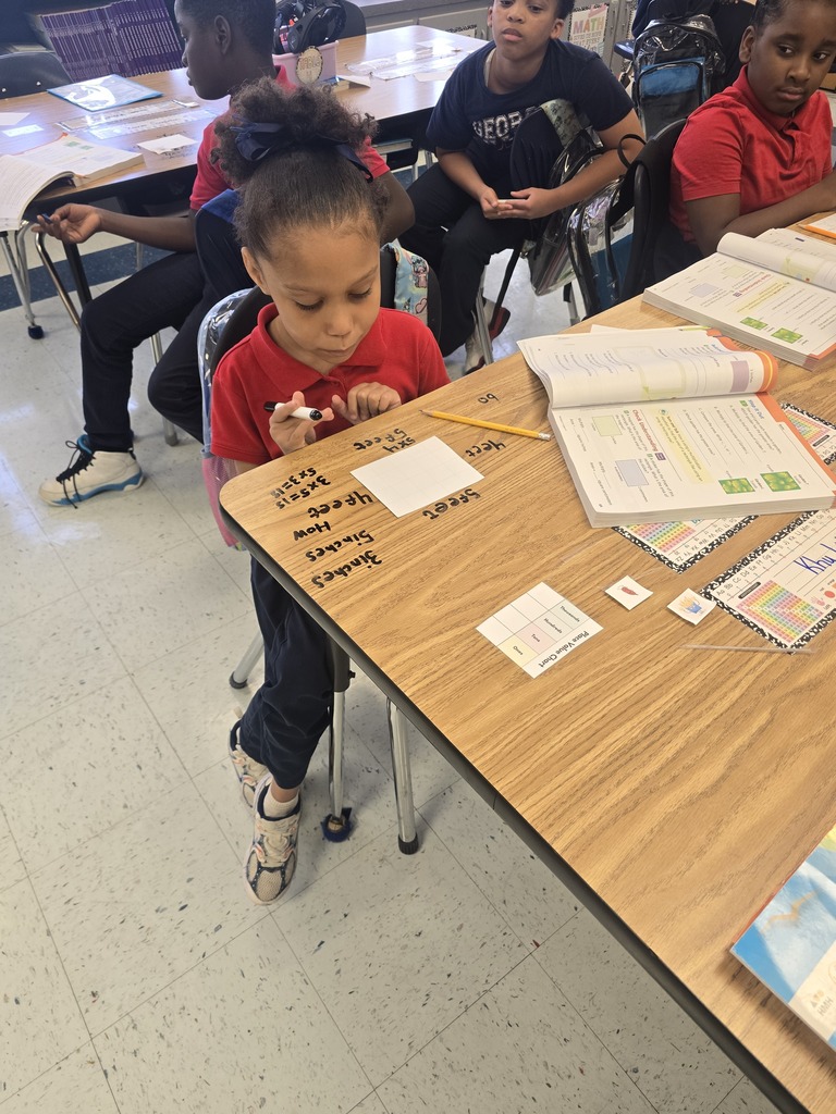 Young children seated at desks, one with a marker writing on paper, others engaged in learning activities.