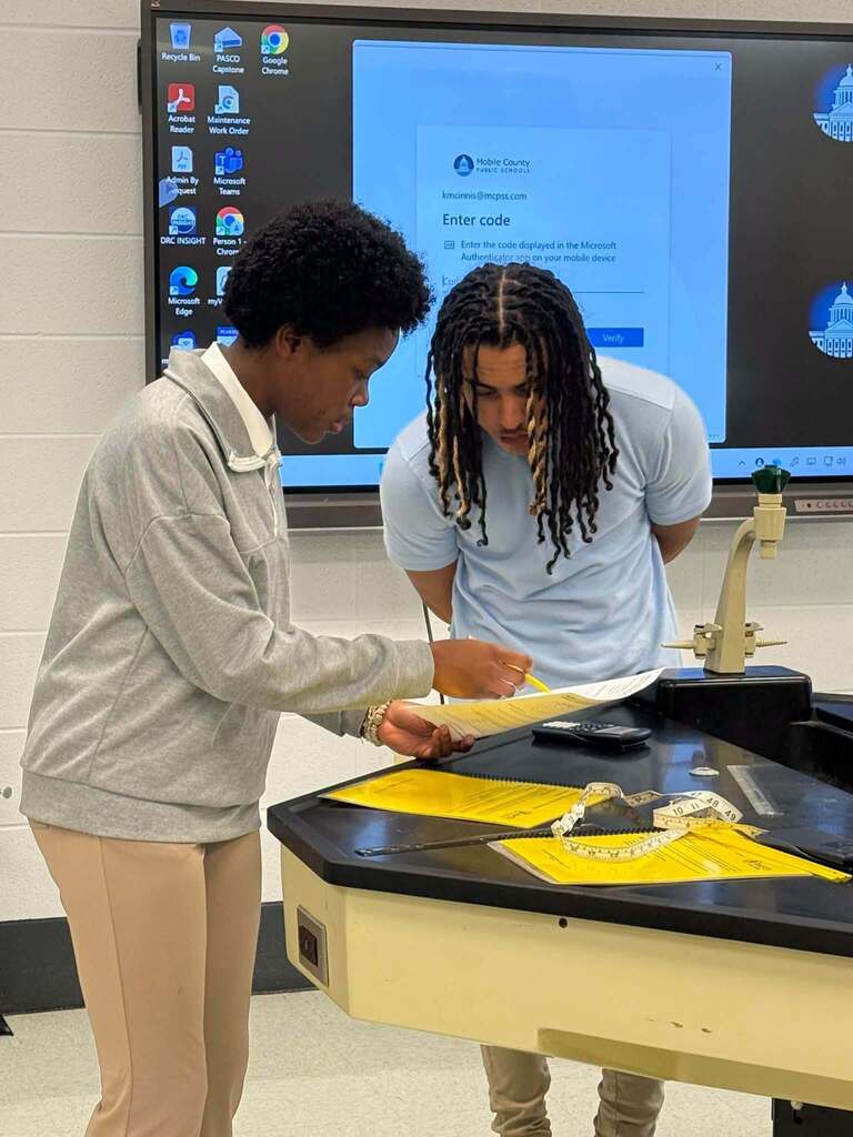 Two people examine a document over a table. A large monitor is in the background.