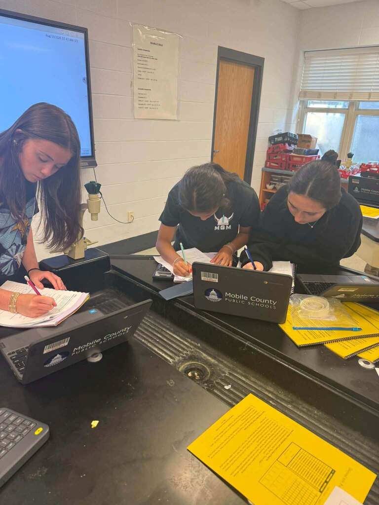 Three students at a table in a room with a monitor and door. Each student has a laptop and papers.
