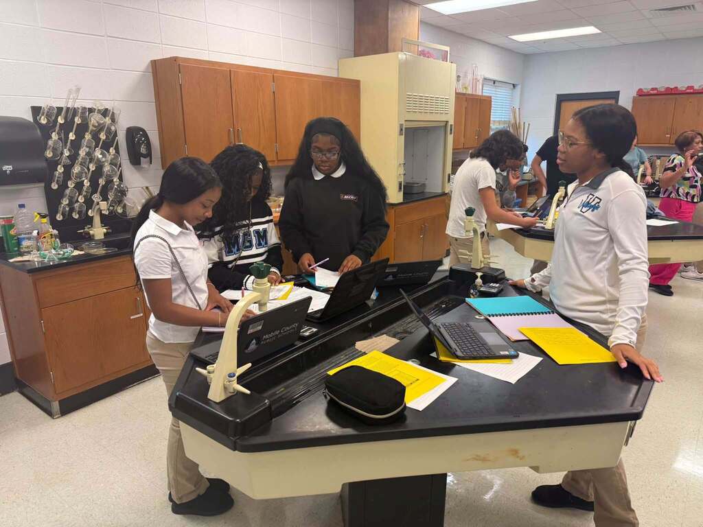 Four students in white shirts and pants work together at a table in a classroom. Behind them, cabinets and shelves are visible.