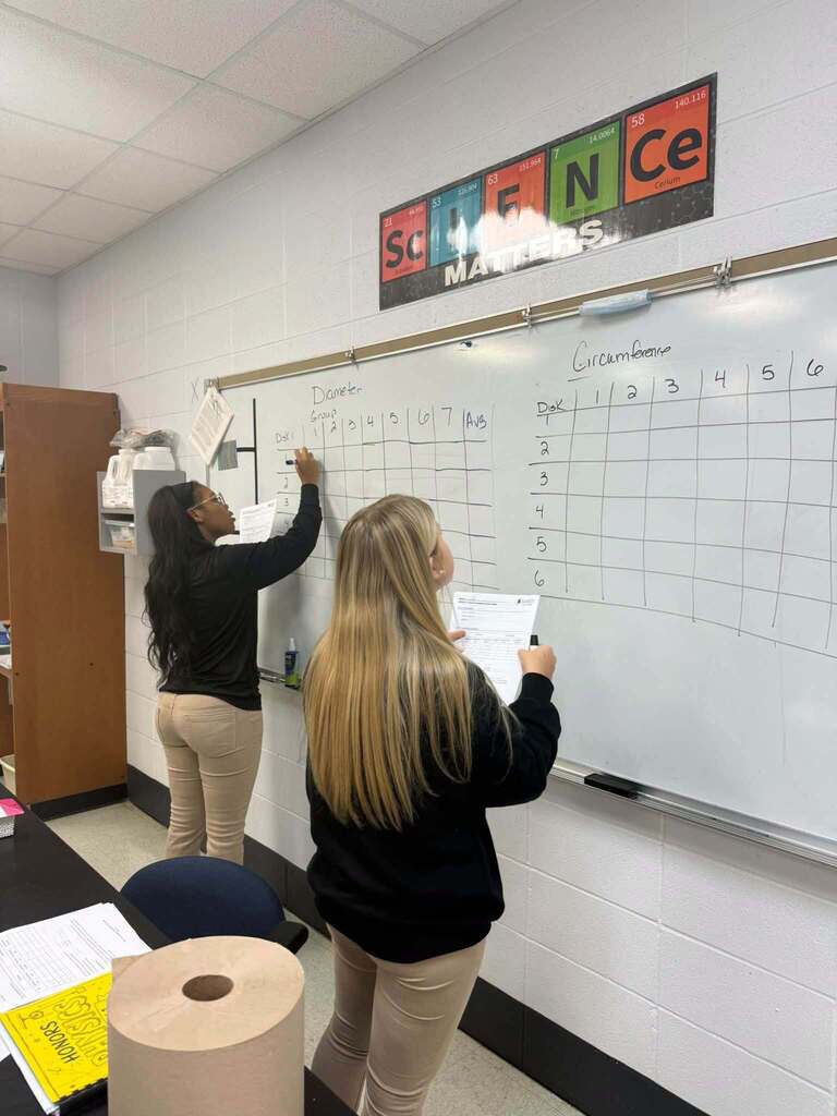 Two students write on a whiteboard in a classroom. One wears a black top, the other a black jacket.