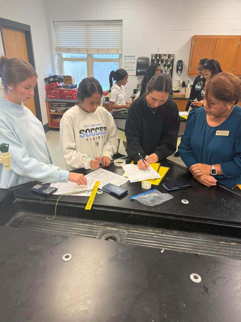 A group of people gathered around a table in a classroom, working on papers and items.