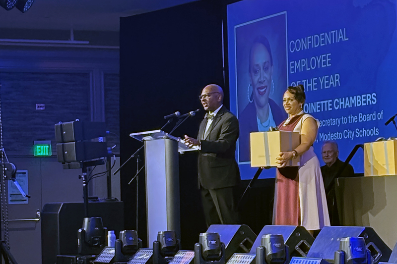 A woman in formal attire holding a box on stage while man at podium is speaking