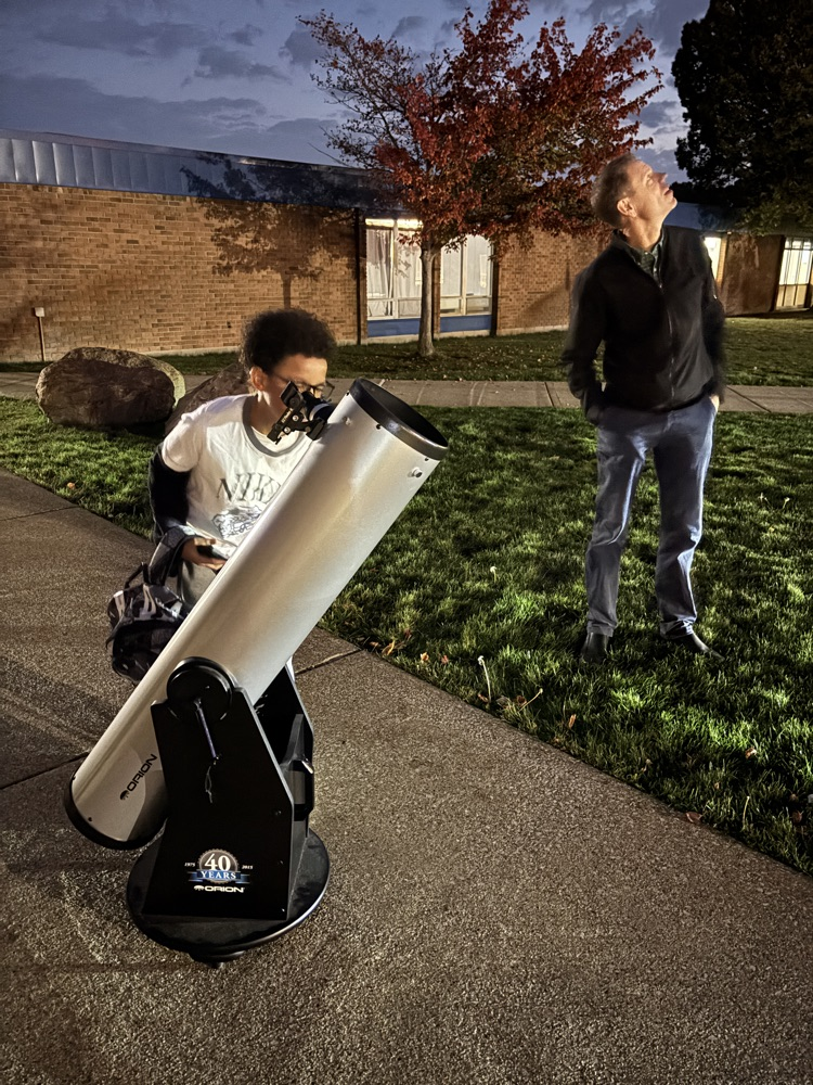 student looking through telescope and teacher beside him looking at the sky