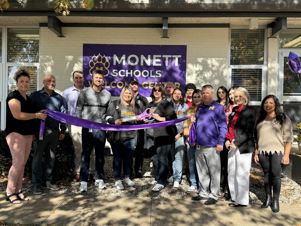 A group of Monett Schools staff and community members stand together outside the new Monett Schools Welcome Center for a ribbon-cutting ceremony. The group is smiling as two people hold large purple scissors and a purple ribbon in front of a building with a Monett Schools Welcome Center banner displayed on the wall.