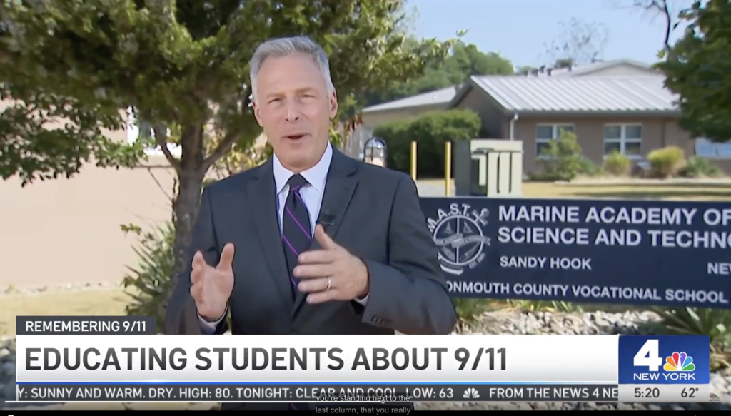 A news reporter in a suit speaks in front of the Marine Academy of Science and Technology at Sandy Hook, part of the Monmouth County Vocational School District. A sign with the school’s name and compass logo is visible beside him. On-screen text reads “Educating Students About 9/11” as part of a News 4 New York segment.