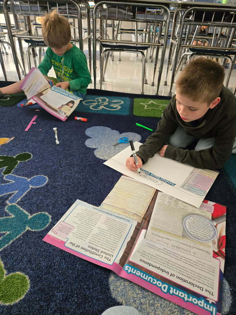 Two third graders sitting on the carpet creating posters using resources on the constitution