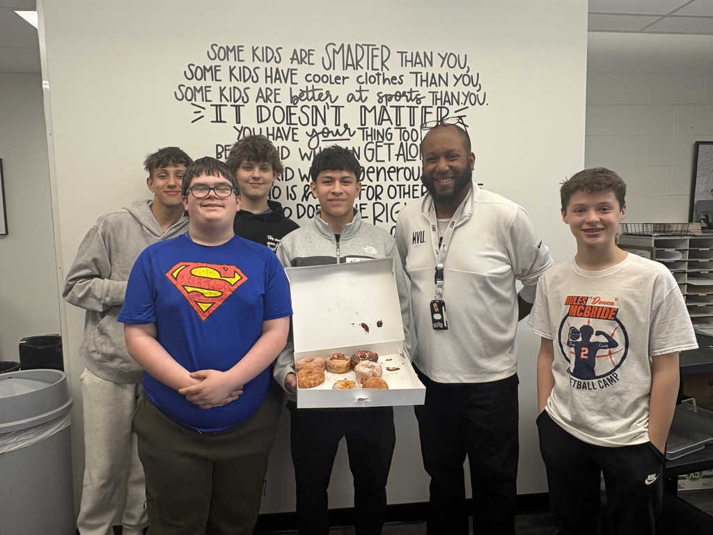 4 students and a teacher. One student is holding an open box of donuts
