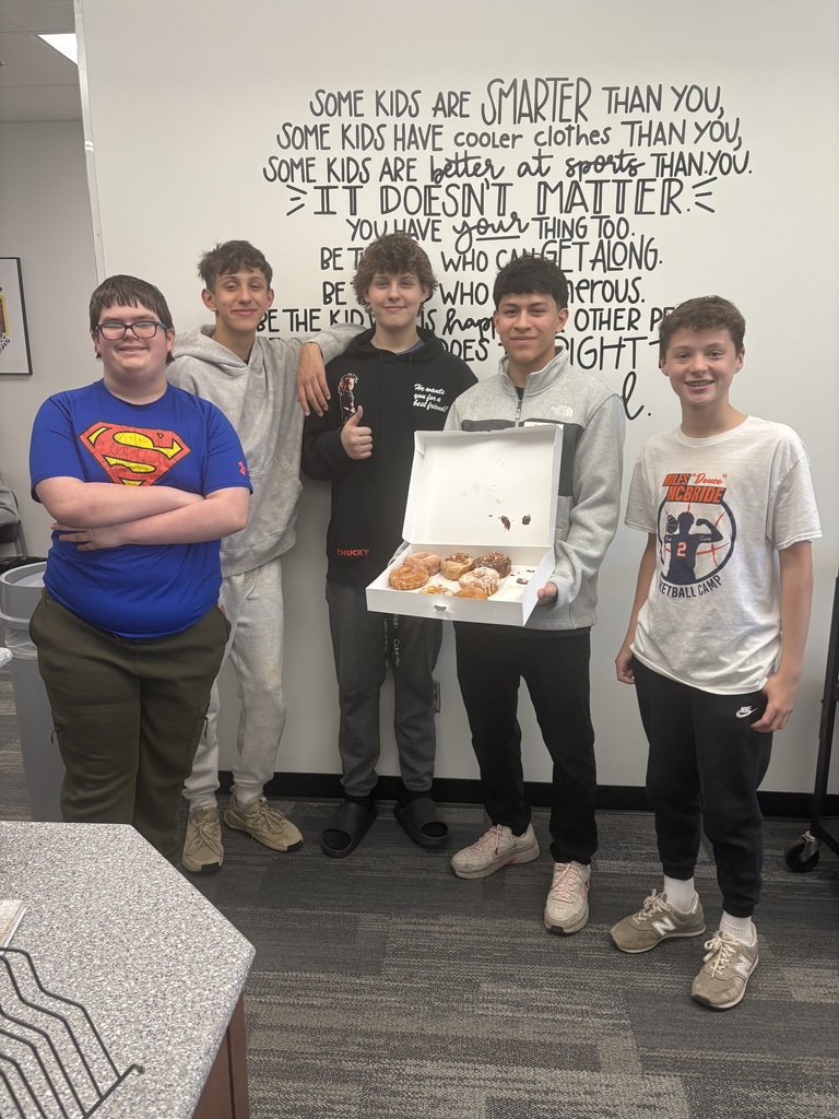 5 students standing in a classroom. 1 of them is holding an open box of donuts