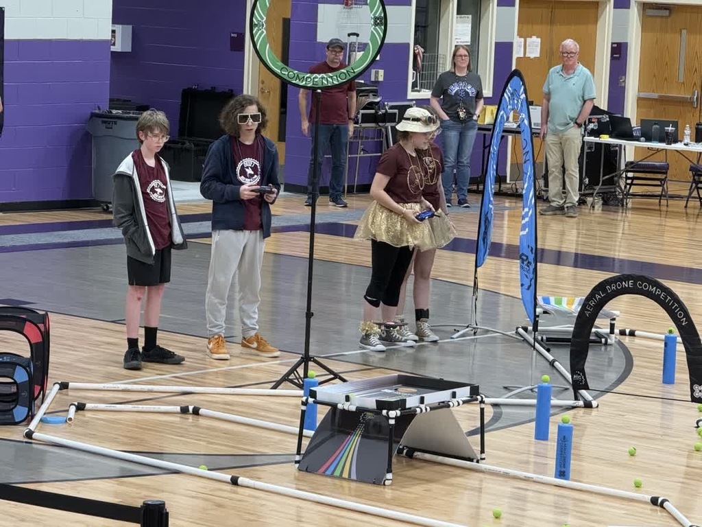 4 students standing in a gym. 2 are holding controllers for a drone and there is an obstacle course. 3 spectators are in the background