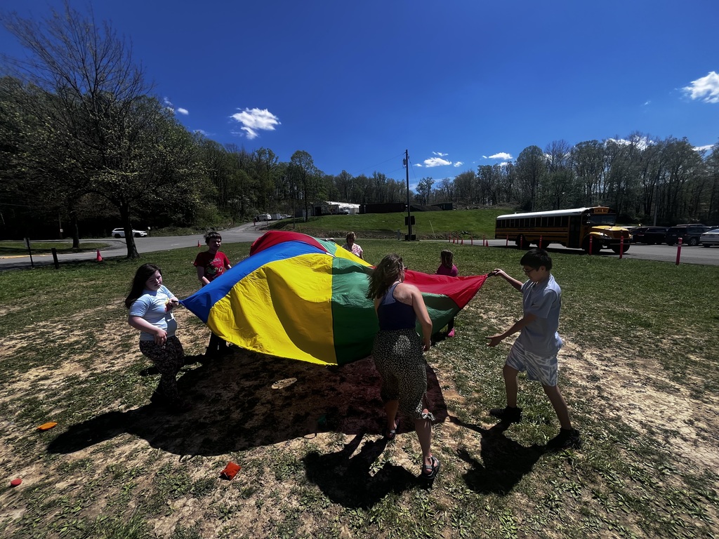 Children stand in a circle outdoors on a sunny day, holding a large multicolored parachute (red, yellow, green, and blue) as it lifts and billows upward. They appear to be playing a group game on a grassy, slightly patchy field. In the background are trees, a road, parked cars, and a yellow school bus. The sky is bright blue with a few small clouds, and strong sunlight creates dark shadows on the ground.