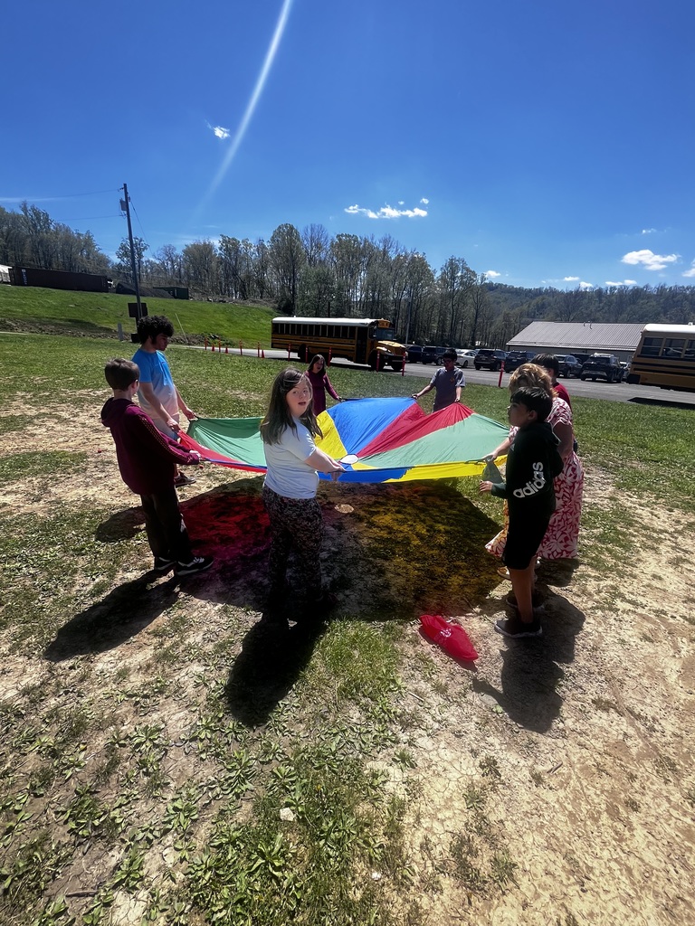 A group of children stands outdoors in a grassy area on a sunny day, holding the edges of a large, colorful parachute (red, blue, green, and yellow). They are spaced in a circle, lifting the parachute slightly. In the background, there are school buses, a parking lot with cars, and a low building. Trees line the horizon under a clear blue sky with a few small clouds. Bright sunlight creates strong shadows on the ground.