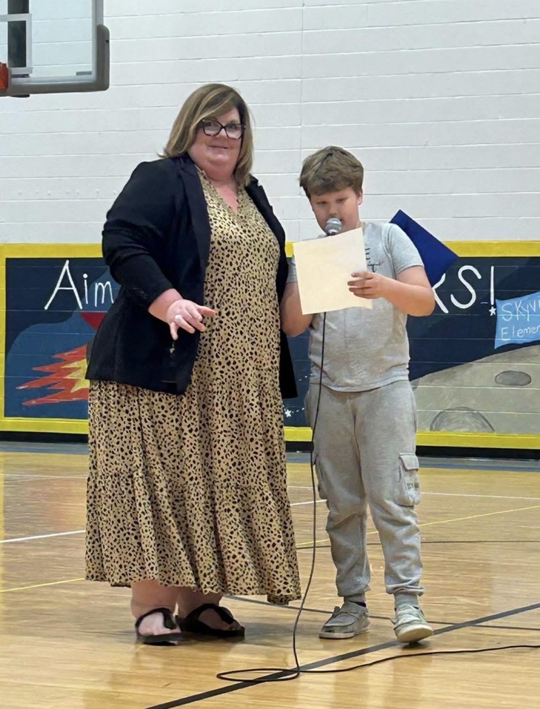 Ms. Cox and a 4th grade boy standing together as he reads his paper about the Source