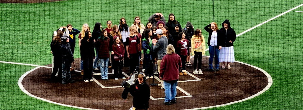 A group of students on a baseball field with a camera man walking away and a choir director standing in front of the students. 