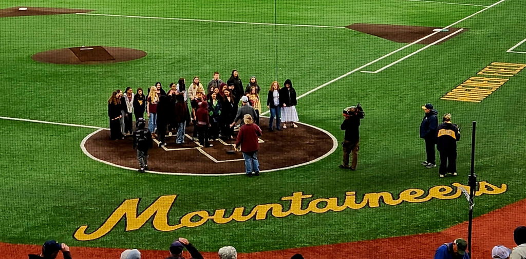 The word Mountaineers is printed on a baseball field and a group of students are standing in the home plate circle. A camera man is holding a camera pointed at the group. There are 2 other people standing on the baseball field.