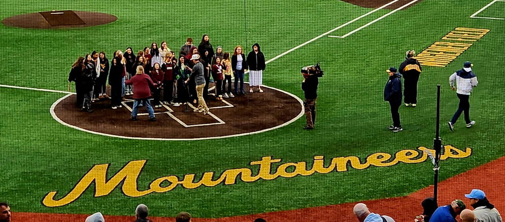 The word Mountaineers is printed on a baseball field with a group of students standing in the home plate circle. A camera man is holding a camera pointed at the group and 3 other people are standing off to the side.