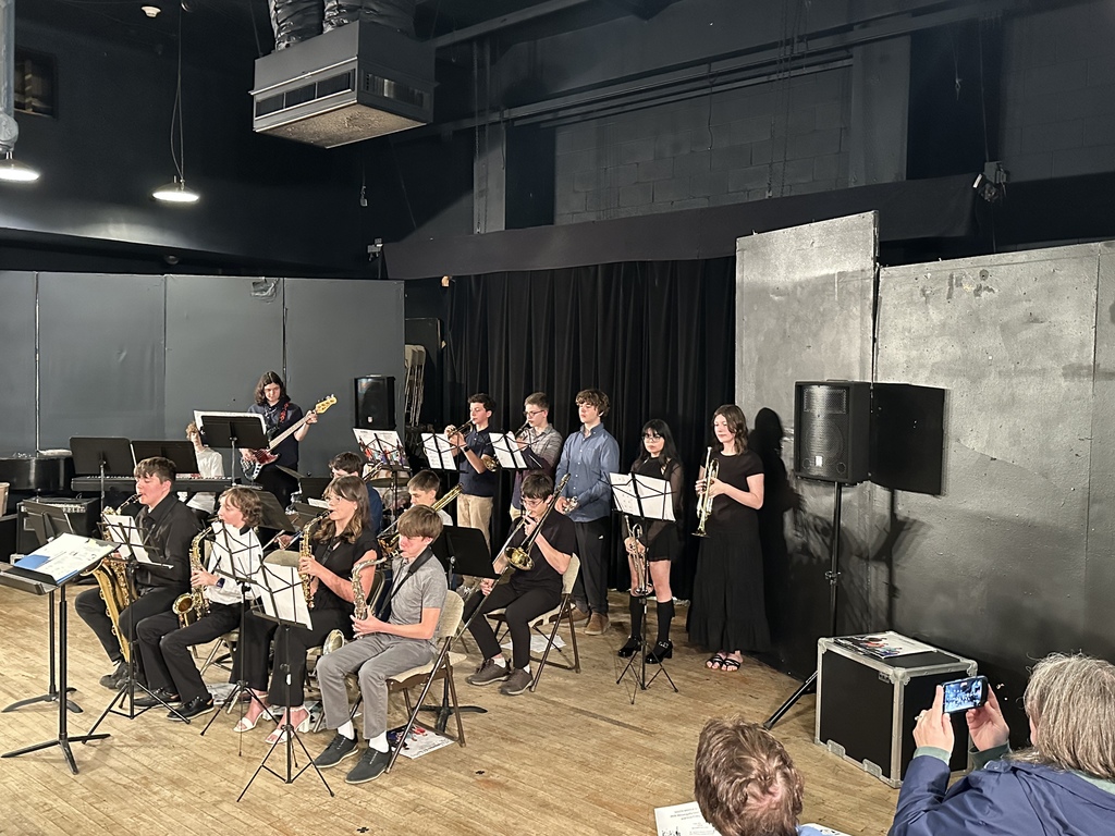 A group of students sitting playing instruments with music stands in front of them. The are in a room with a wooden floor and it has a black ceiling.