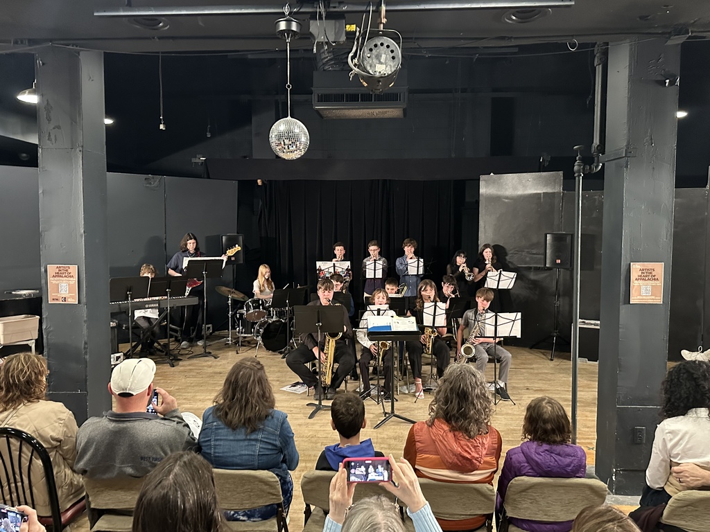 A group of students sitting playing instruments with music stands. There are people sitting in folding chairs watching the students perform. The room  has gray walls and a black ceiling.
