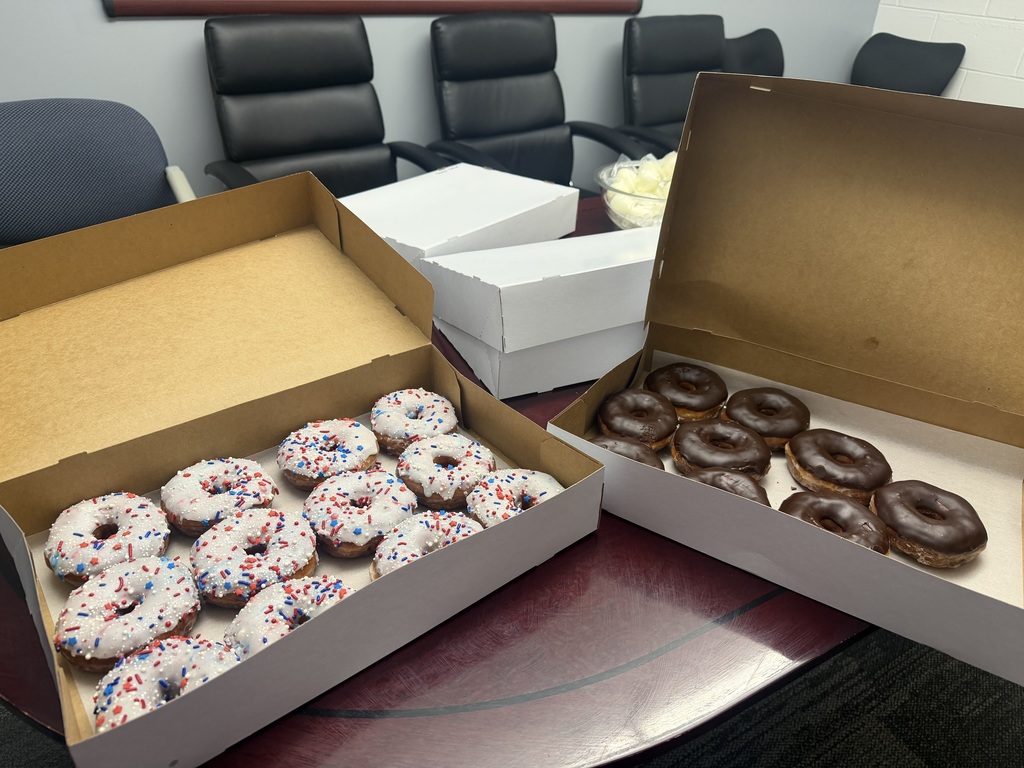 There are 2 open boxes of donuts. One box has 12 white iced donuts with rainbow sprinkles on them and the other opened box has 9 chocolate iced donuts in it. There are 2 closed boxes on a table. In the background you can see 6 chairs against the wall.