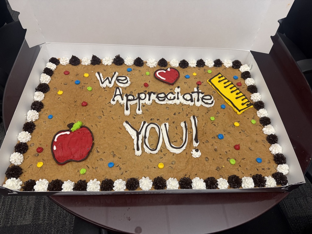 A Square Chocolate Chip Cookie with an apple, the words we appreciate you, a heart, and a ruler on it. The cookie has a black and white icing border and is laying in an open box that is sitting on a table.