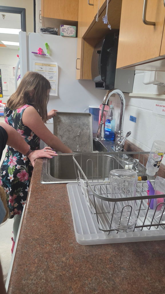 Girl in a flowered outfit standing at a sink washing a cookie sheet. 
