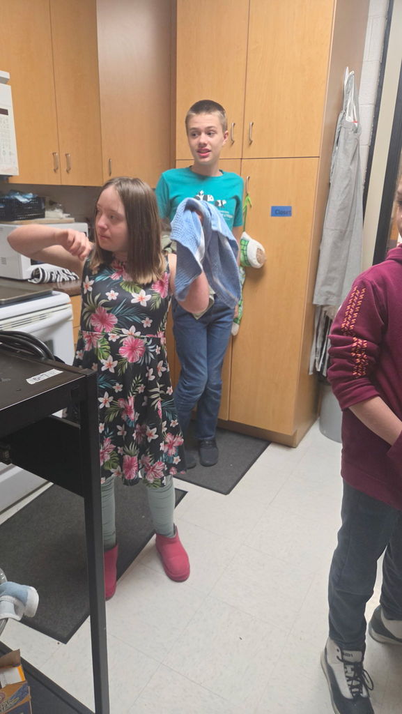 A girl is standing in a room holding a dish towel. There is a boy behind her.