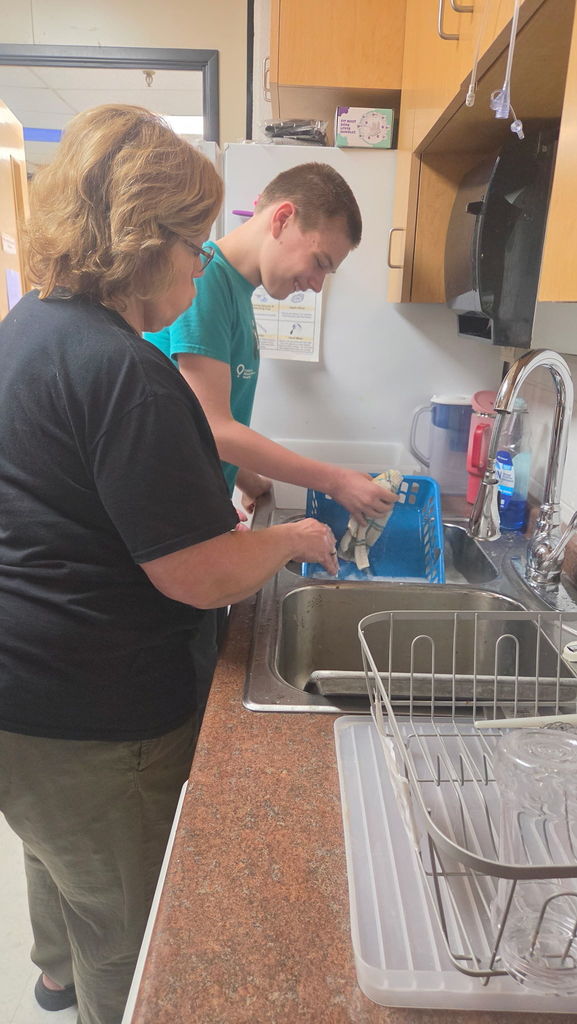 A boy is scrubbing a basket at the sink with a dish rag. A woman is standing beside him at the sink. They are in a kitchen.