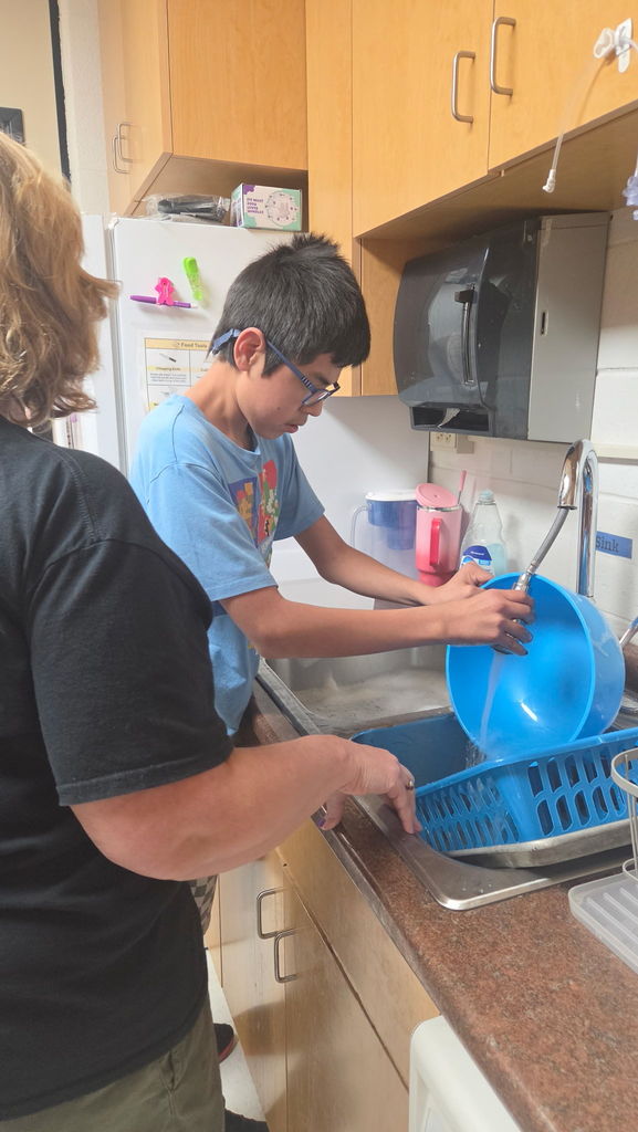 A boy at the sink running water over a bowl. A woman is holding a basket at the sink.