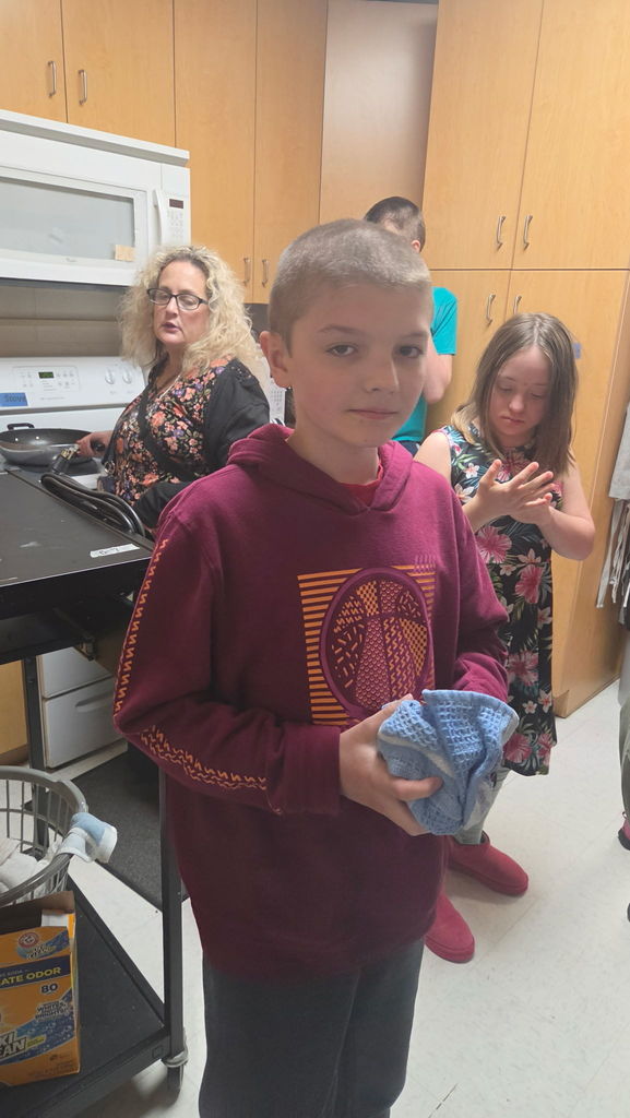 A boy holding a tea towel. There is a woman and young girl in the background.