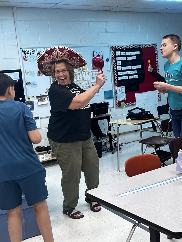 Woman holding a shaker and wearing a sombrero. She is in a classroom with 2 boys looking a her.
