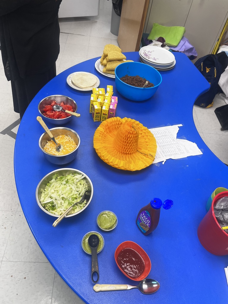 There is a blue table with salsa, lettuce, cheese, tomatoes, juice boxes, beef, and taco shells. There is a small sombrero as a decoration in the middle of the table.