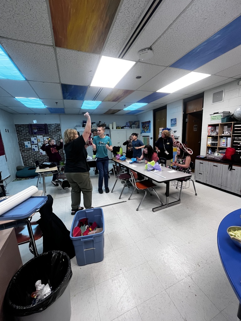 A woman with her hands up is standing in front of a table. There are 5 students looking at the woman. They are in a classroom.