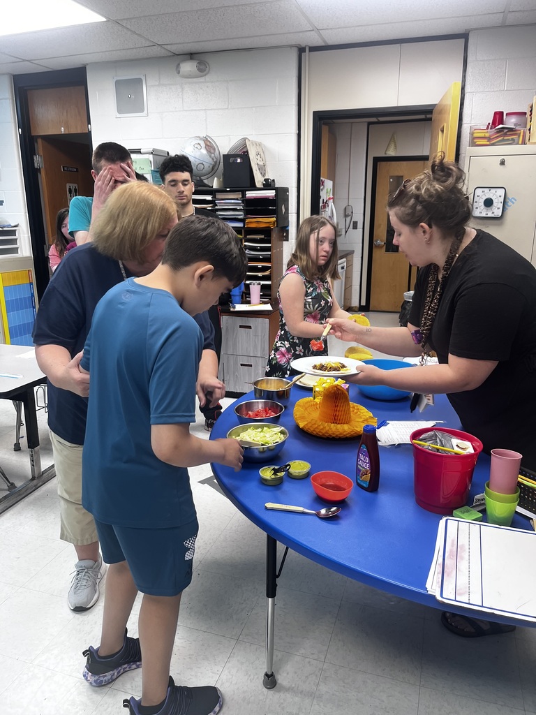 4 students and a woman are standing around a blue table with food on it. Another woman is standing behind the table putting food on a white plate. They are all in a classroom.