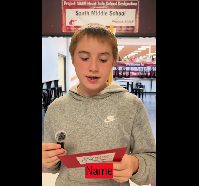 Student in grey sweatshirt reading from a red card.