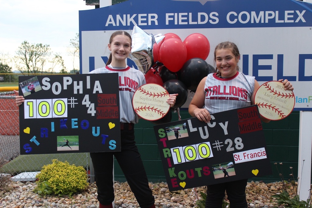 Two softball girls holding up posters and softball signs. There are red and black balloons in the background along with an Anker Fields Complex blue and white sign. There is a flower bed with a bush and rocks in the background.