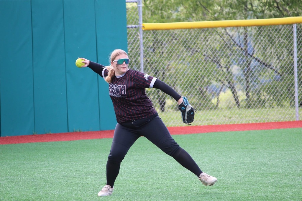 A softball player is throwing a yellow softball. She has sunglasses on and is wearing a black mitt. There is a part of a blue building and fence in the background.