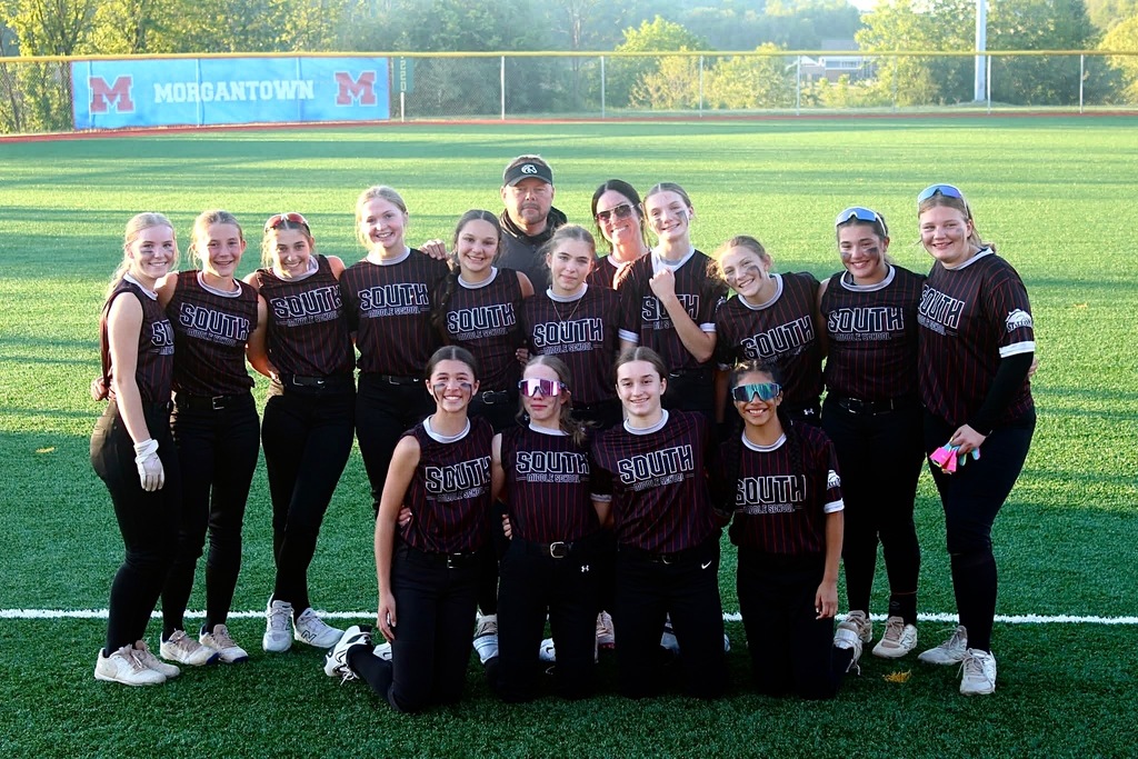 This is a team South photo. 4 girls are on their knees and 10 girls are standing behind them. There is a man and a woman behind the girls standing. There is a fence with a Morgantown banner in the background.