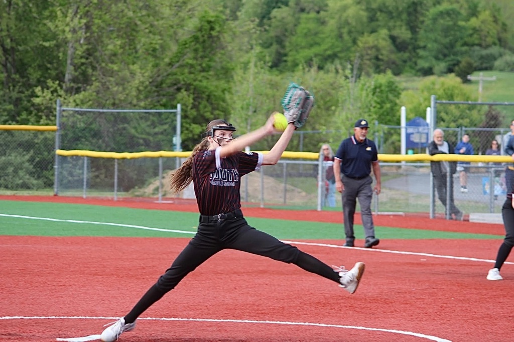 A picture is getting ready to throw a yellow softball. An umpire is walking a baseline and spectators are standing behind a fence in the background.