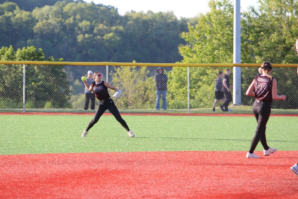 A softball player wearing a mitt and sunglasses is getting ready to throw a yellow softball. There is another player looking at the girl with the ball and spectators are standing behind a fence in the background.