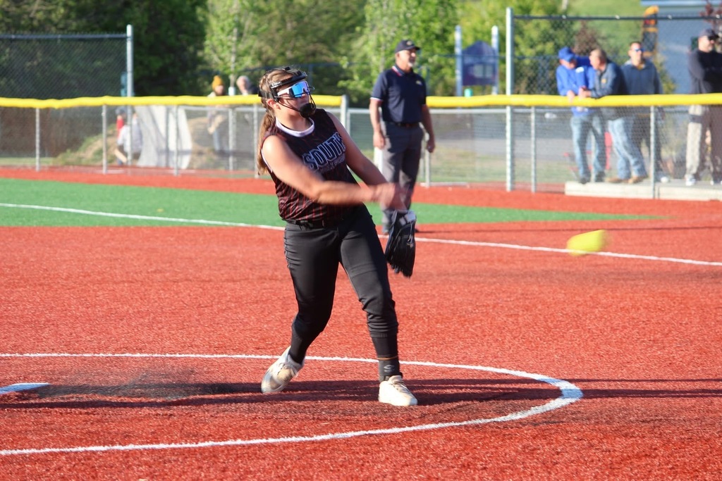 Softball player pitching a ball. She has sunglasses and a mask on. There is an umpire standing behind her. In the background there is a fence, trees, and spectators.