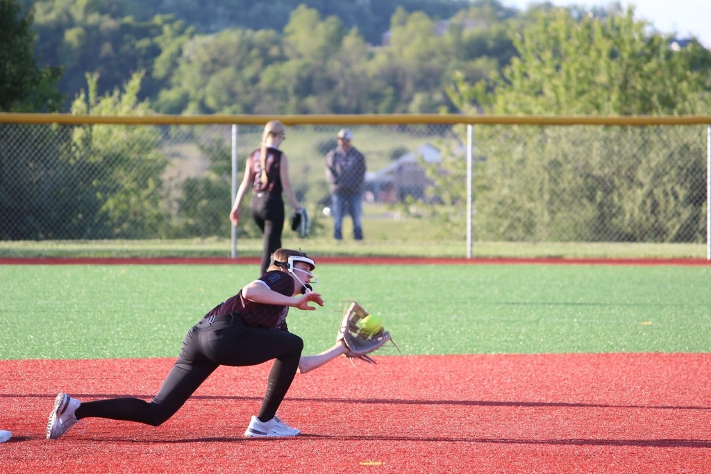 A softball player is bent down and has a yellow softball in her mitt. In the background another player is walking towards a fence. In the background there is a fence, a man standing behind the fence and trees.
