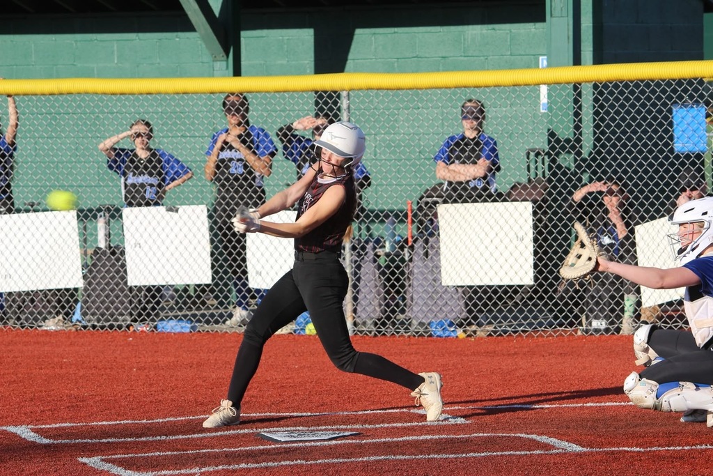 A softball player swinging a bat. You can see a yellow softball has been hit and a catcher is holding her mitt up. In the background there is a fence and player are watching behind the fence.