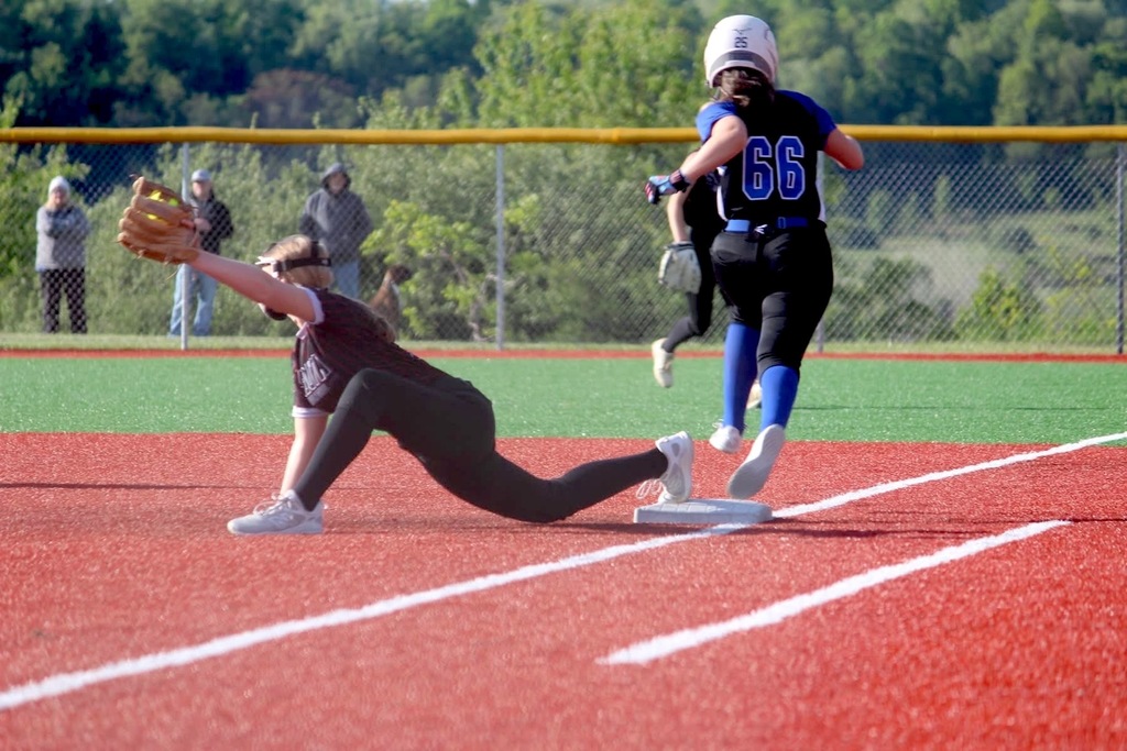 A softball player has caught a yellow softball while having a foot on a base and a player wearing #66 on her jersey has run past the base. In the background there is a fence and 3 spectators are standing behind the fence.