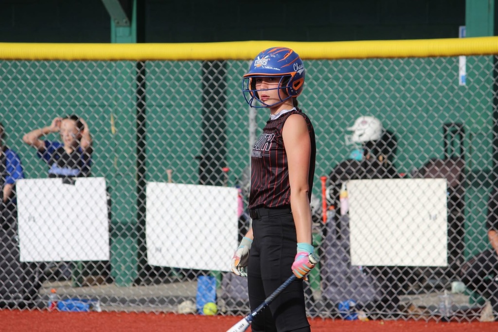 A softball player wearing a blue and orange helmet is holding a bat. There is a fence and softball players in the background.