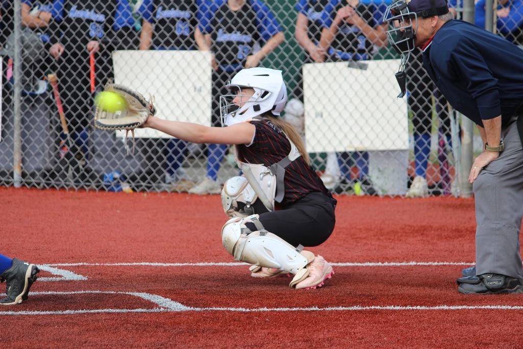 A catcher is catching a yellow softball in her mitt. An umpire is standing behind her. There is a fence with players wearing blue uniforms in the background.