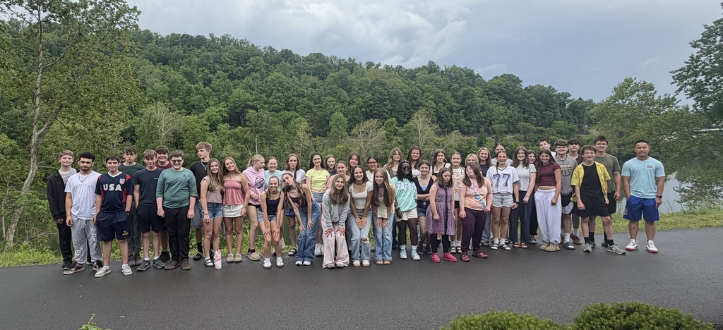 A  group of students stand along a paved trail. There are trees in the background.