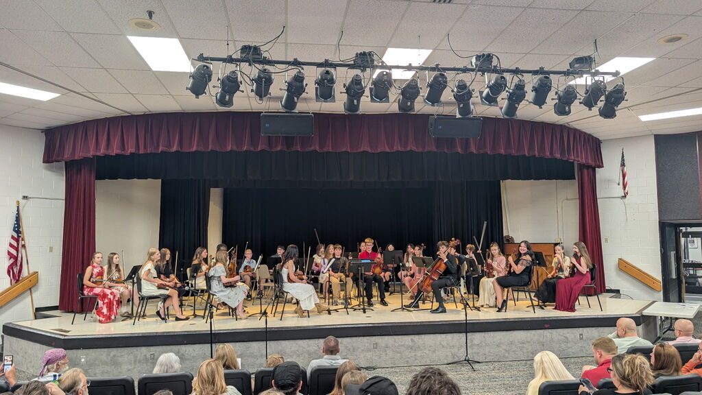 A group of students sitting on a stage in an auditorium holding instruments. There are people sitting across from them as an audience. 
